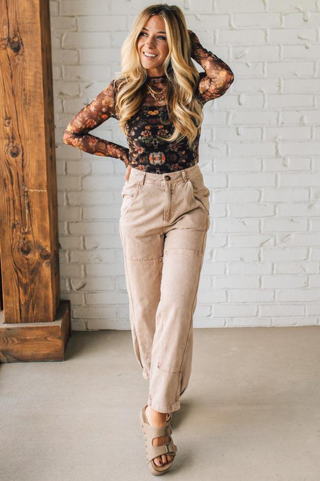 Woman wearing a black lace top and beige pants standing against a white brick wall.