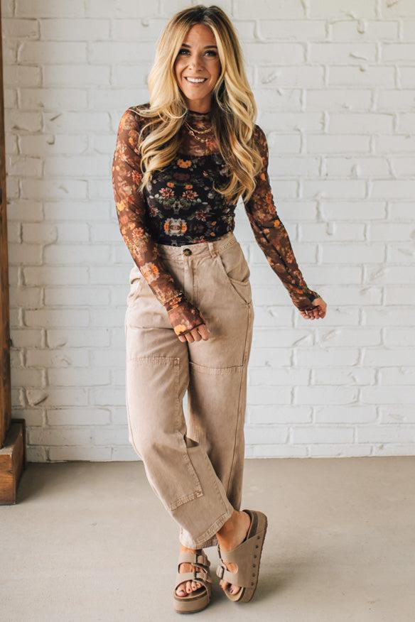 Woman wearing a floral long-sleeve top and beige pants against a white brick wall.