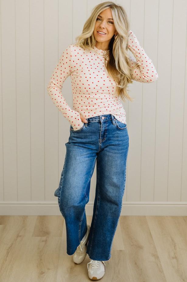 Woman wearing a polka dot blouse and blue jeans standing against a light wooden paneled wall.
