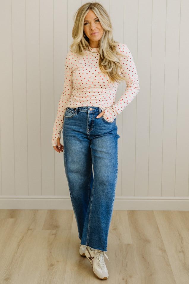 Woman wearing a polka dot blouse and blue jeans standing against a neutral wall.