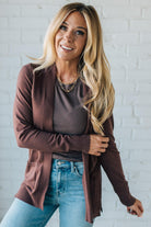 Woman wearing a brown cardigan over a gray shirt with blue jeans against a white brick wall.