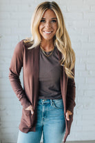 Woman wearing a brown cardigan, brown shirt, and blue jeans against a white brick wall.