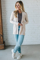 Woman wearing a beige cardigan, gray top, light blue jeans, and white sneakers against a white brick wall.