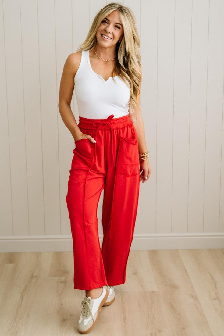 Woman wearing a white tank top and red pants standing against a light wooden paneled wall.