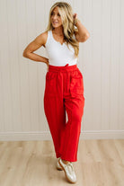 Woman wearing a white top and red pants standing against a light wooden paneled wall.