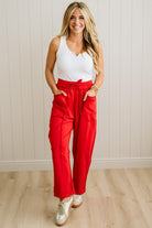 Woman wearing a white top and red pants standing against a light wooden paneled wall.