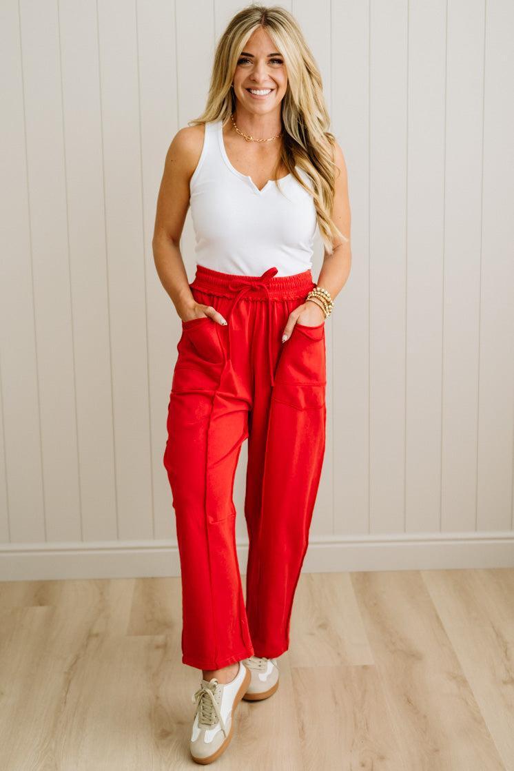 Woman wearing a white top and red pants standing against a light wooden paneled wall.