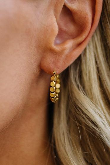 A close-up of a model's ear wearing a small gold hoop earring with textured, beaded segments.