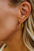 A close-up of a woman's ear wearing a small gold hoop earring with textured, beaded segments.