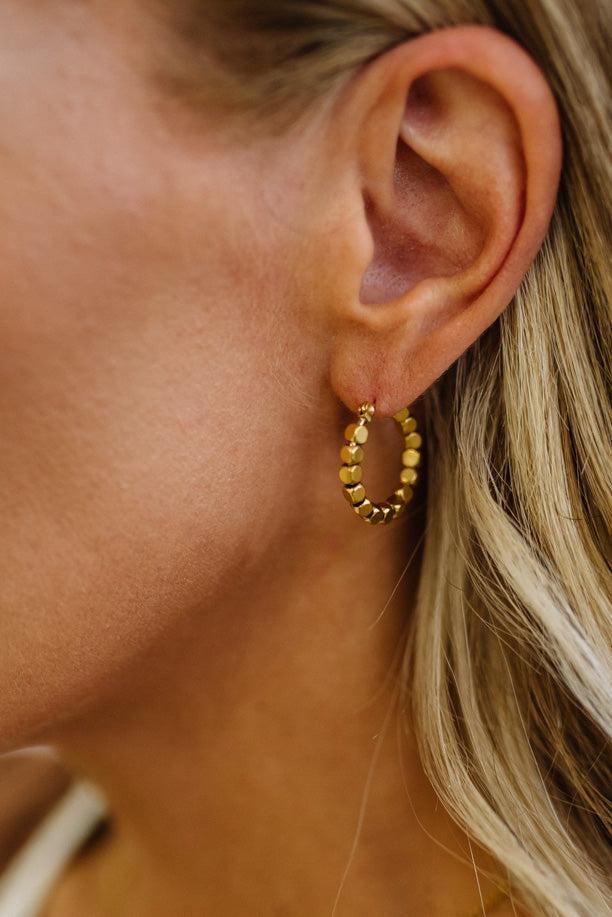 A close-up of a woman's ear wearing a small gold hoop earring with textured, beaded segments.