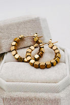 A pair of chunky gold hoop earrings with faceted, cube-like beads, resting on a textured beige jewelry box.