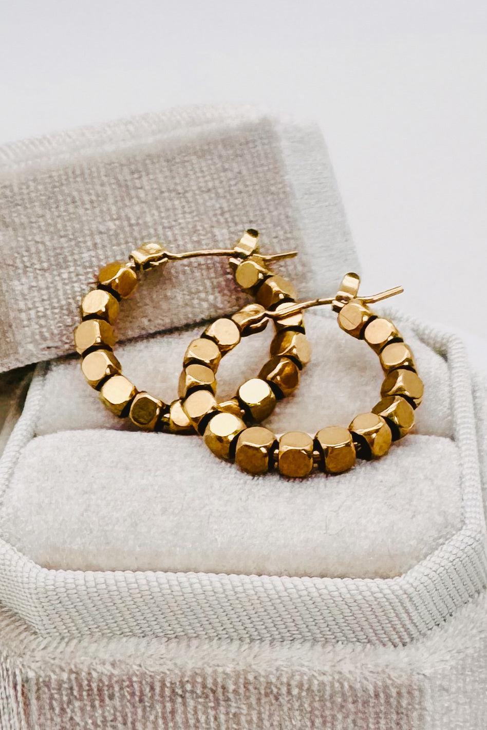 A pair of chunky gold hoop earrings with faceted, cube-like beads, resting on a textured beige jewelry box.