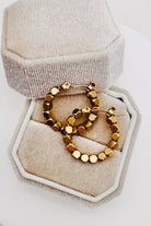 A pair of gold hoop earrings with faceted round beads, displayed inside an open hexagonal beige velvet jewelry box on a white surface.