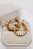 A pair of gold hoop earrings with faceted round beads, displayed inside a textured beige jewelry box.