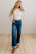 A woman modeling a beige long-sleeve crewneck top and dark blue wide-leg cuffed jeans with brown suede boots, standing in a room with light wood floors and white paneled walls.
