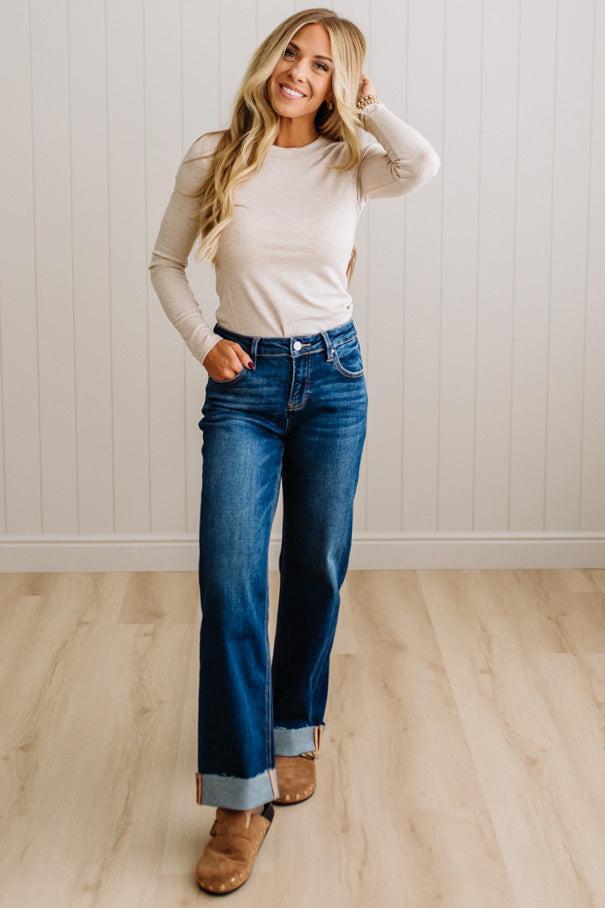 A woman modeling a beige long-sleeve crewneck top and dark blue wide-leg cuffed jeans with brown suede boots, standing in a room with light wood floors and white paneled walls.