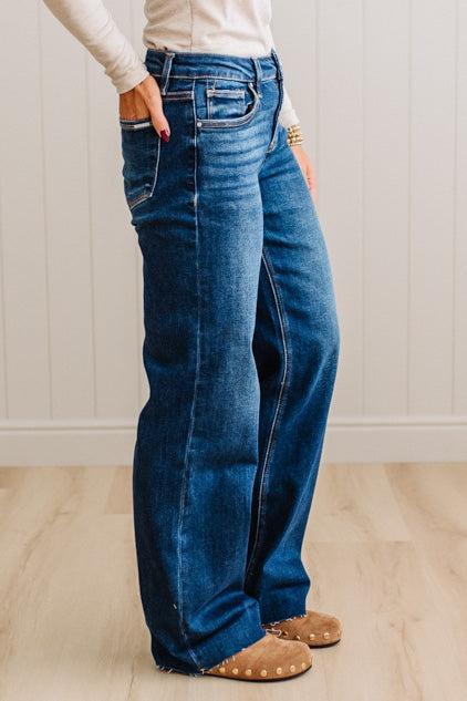 A person wearing dark blue wide-leg denim jeans with a cream long-sleeve top and tan suede shoes, standing in a room with light wood floors and white paneled walls.