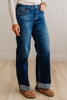 A person wearing dark blue straight-leg denim jeans with rolled cuffs, paired with a cream long-sleeve top and brown suede slippers, standing on a light wood floor against a white paneled wall.
