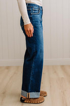 A person wearing dark blue wide-leg denim jeans with a rolled cuff, paired with a cream long-sleeve top and brown suede slippers, standing on a light wood floor against a white paneled wall.
