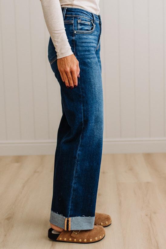 A person wearing dark blue wide-leg denim jeans with a rolled cuff, paired with a cream long-sleeve top and brown suede slippers, standing on a light wood floor against a white paneled wall.