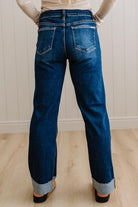 A person shown from the back wearing dark blue straight-leg jeans with rolled cuffs, standing on a light wood floor against a white paneled wall.