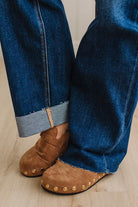 Close-up of a person wearing blue jeans with cuffed hems and brown suede slip-on shoes with gold-tone studs, standing on a light wood floor.