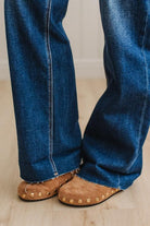 Close-up of a person wearing blue denim jeans with frayed hems and tan suede slip-on shoes with brass studs, standing on a light wood floor.