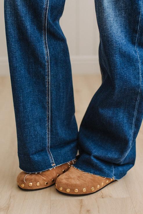 Close-up of a person wearing blue denim jeans with frayed hems and tan suede slip-on shoes with brass studs, standing on a light wood floor.
