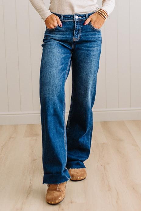 A person wearing blue wide-leg jeans with a cream long-sleeve top and tan suede boots, standing on a light wood floor against a white paneled wall.