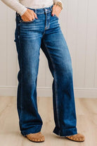 A person wearing dark blue wide-leg denim jeans with a white long-sleeve top and tan slip-on shoes, standing against a white paneled wall and light wood floor.
