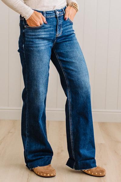 A person wearing dark blue wide-leg denim jeans with a white long-sleeve top and tan slip-on shoes, standing against a white paneled wall and light wood floor.