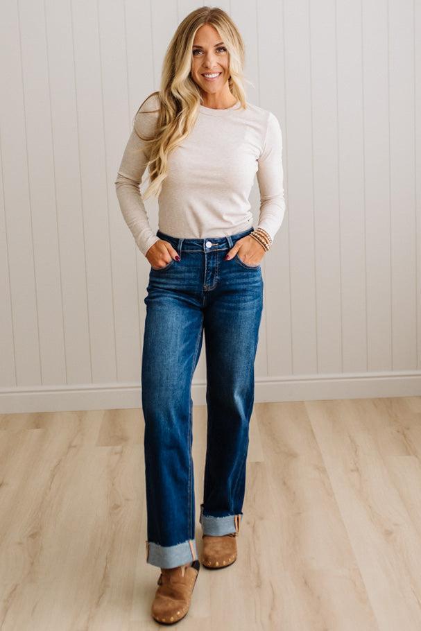 A woman modeling a cream long-sleeve knit top and dark blue straight-leg jeans with rolled cuffs, paired with brown suede boots, standing in front of a white paneled wall and light wood floor.