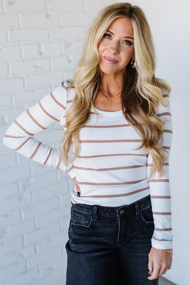 Woman wearing a striped long-sleeve top and dark jeans against a white brick wall.