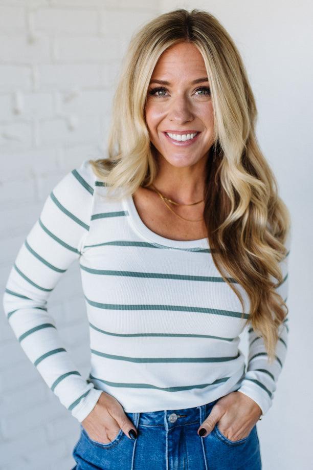 Woman wearing a white and green striped long-sleeve top with blue jeans against a light gray background