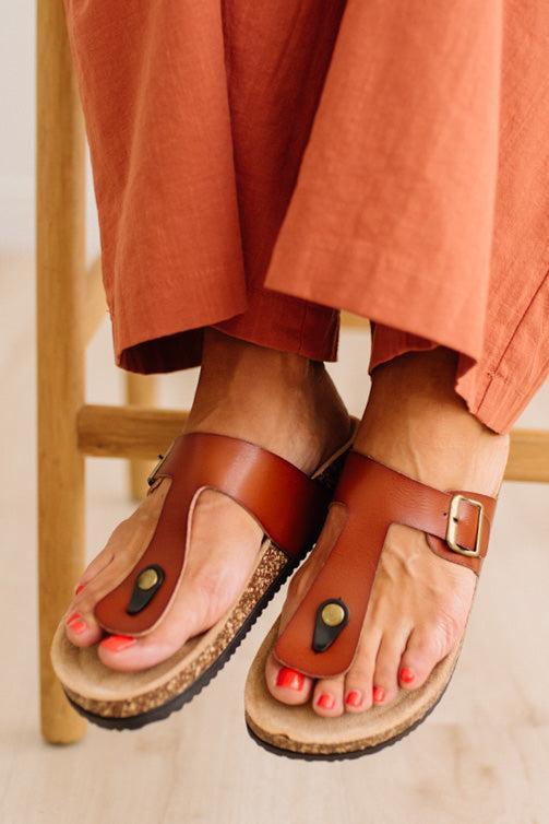A close-up of a person wearing brown leather T-strap sandals with cork soles and red toenail polish, with rust-colored pants visible against a light wood chair and floor.