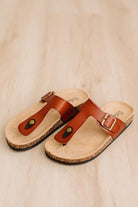 A pair of brown leather thong sandals with cork soles and gold-tone buckles, displayed on a light wood floor.