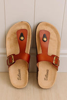 A pair of Birkenstock Bellemanie slide sandals with tan cork footbeds and reddish-brown leather straps, displayed on a light wood floor against a white paneled wall.