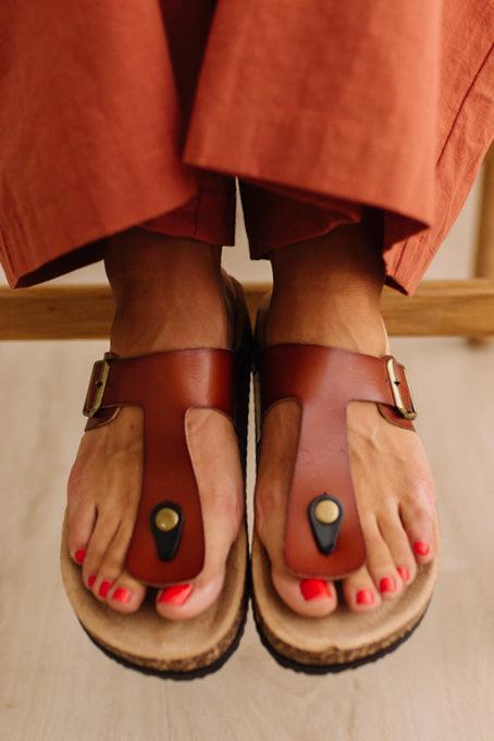 A close-up of a person wearing brown leather T-strap sandals with cork-look soles, paired with rust-orange pants. The sandals feature a decorative black and gold accent on the toes.