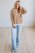 A woman modeling a brown and cream striped long-sleeve henley sweater with light-wash blue flare jeans, standing in a room with light wood floors and white paneled walls.
