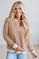 A smiling blonde woman wearing a brown and cream horizontally striped knit sweater with a buttoned henley neckline, paired with light blue jeans, standing against a light-colored wall.