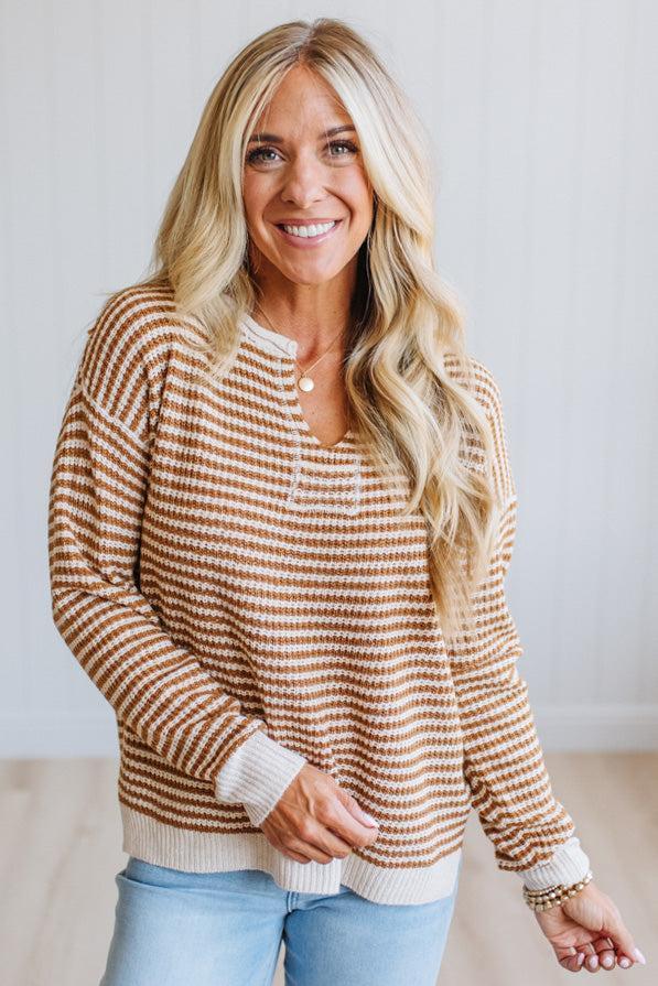 A smiling blonde woman wearing a brown and cream horizontally striped knit sweater with a buttoned henley neckline, paired with light blue jeans, standing against a light-colored wall.