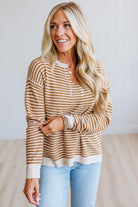 A smiling blonde woman wearing a brown and cream striped knit sweater with a henley-style neckline, paired with light blue jeans, standing in a bright indoor setting.