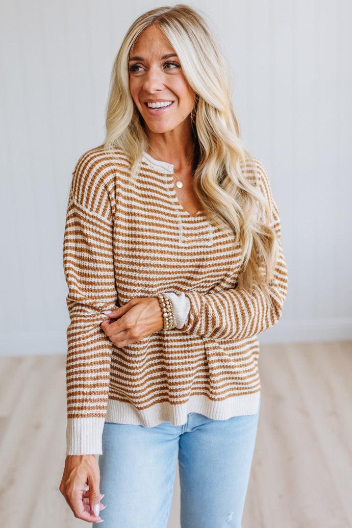 A smiling blonde woman wearing a brown and cream striped knit sweater with a henley-style neckline, paired with light blue jeans, standing in a bright indoor setting.