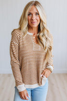A blonde woman wearing a brown and cream horizontally striped waffle-knit sweater with a henley neckline, paired with light blue jeans, standing against a white paneled wall.