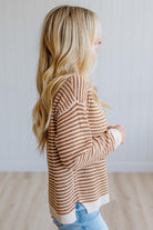 A woman with long blonde hair is shown from the side profile wearing a brown and cream striped knit sweater and light blue jeans, standing in a brightly lit room with a white paneled wall and light wood floor.