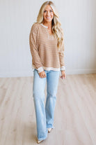 A woman modeling a brown and cream striped long-sleeve henley top with light blue jeans, standing in a bright room with light wood floors and white paneled walls.