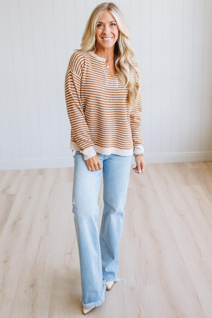 A woman modeling a brown and cream striped long-sleeve henley top with light blue jeans, standing in a bright room with light wood floors and white paneled walls.