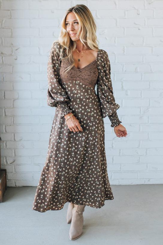 Woman wearing a brown floral dress against a white brick wall.