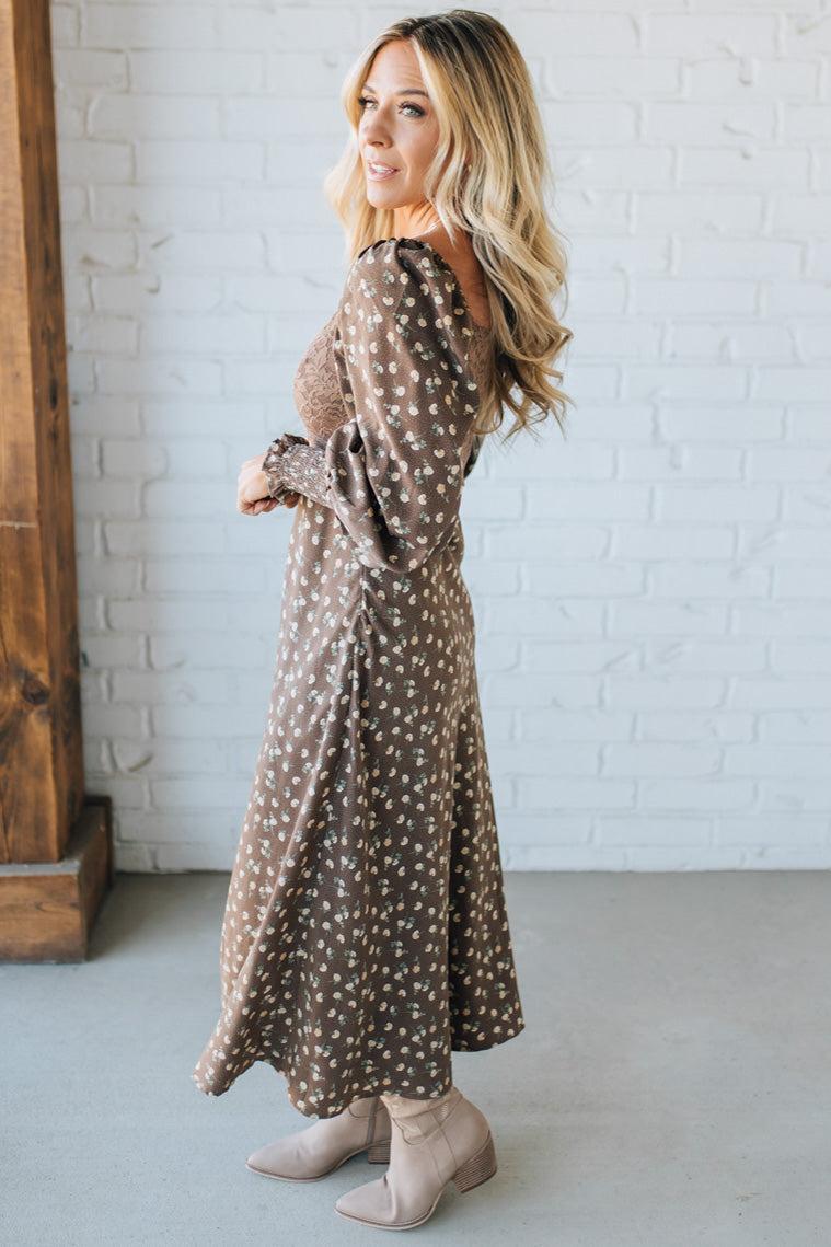 Woman wearing a long, patterned dress standing against a white brick wall.