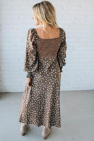 Woman wearing a brown patterned dress against a white brick wall.